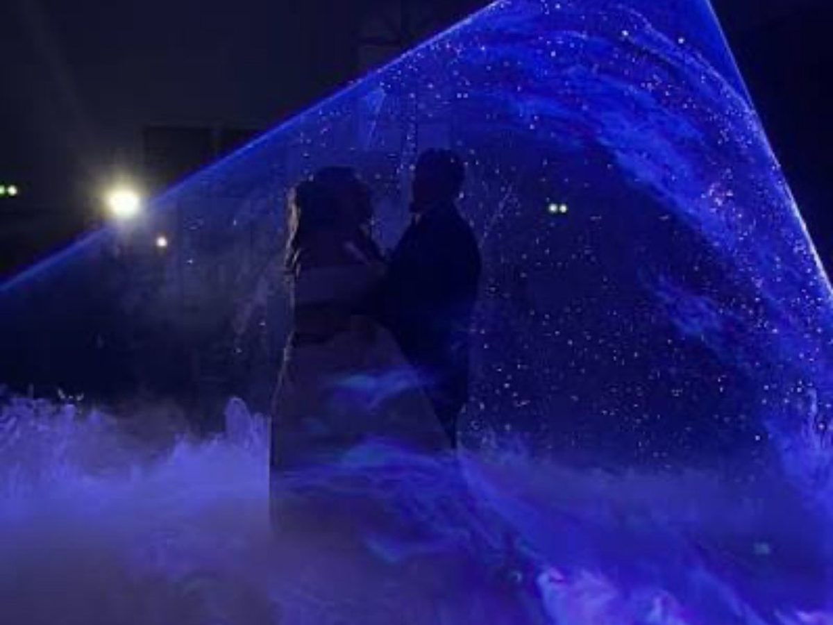 A newly married couple embracing at the centre of a cone of blue light, on a carpet of fog across the floor.